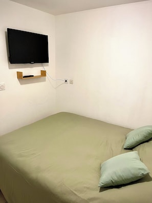 Ein Einzelbett mit grüner Matratze, ein an der Wand befestigter Fernseher und eine Steckdose.