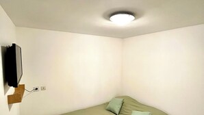 A small, single-bed room with a green bedspread, a mounted television, and a wooden nightstand. - Cosy, modern studio - Gard (Saint-Ambroix)
