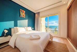 Ein Hotelzimmer mit einem Bett, einem Nachttisch, einer Lampe, einem Gemälde und einem großen Fenster mit Blick aufs Meer.