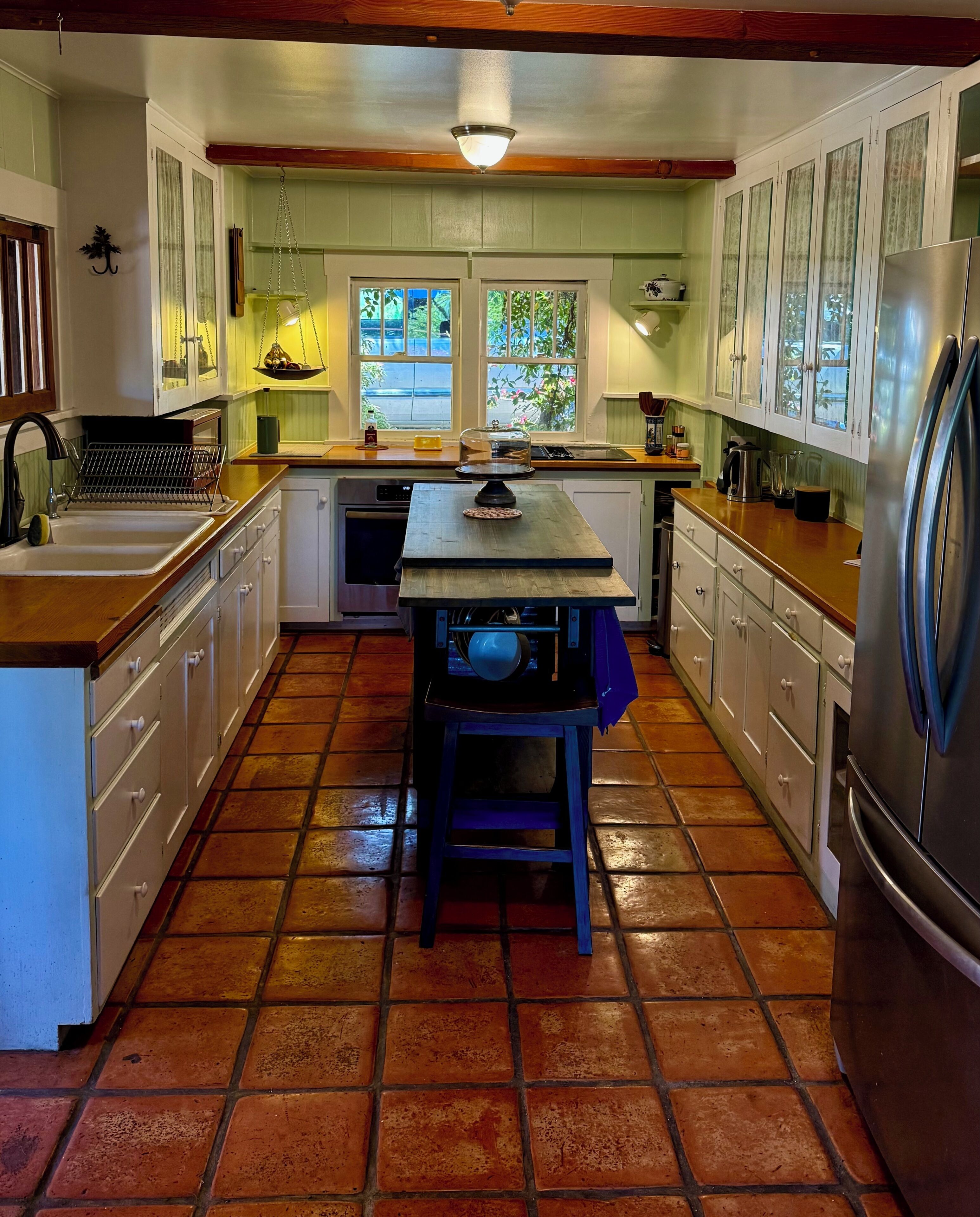 Una cocina con mesa de madera, armarios blancos, piso de baldosas y una lámpara colgante.