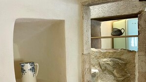 Uma escadaria de pedra leva a um nível superior, uma parede branca com uma pequena prateleira exibindo uma caneca decorativa, e um vislumbre de um quarto com um armário azul claro ao fundo.