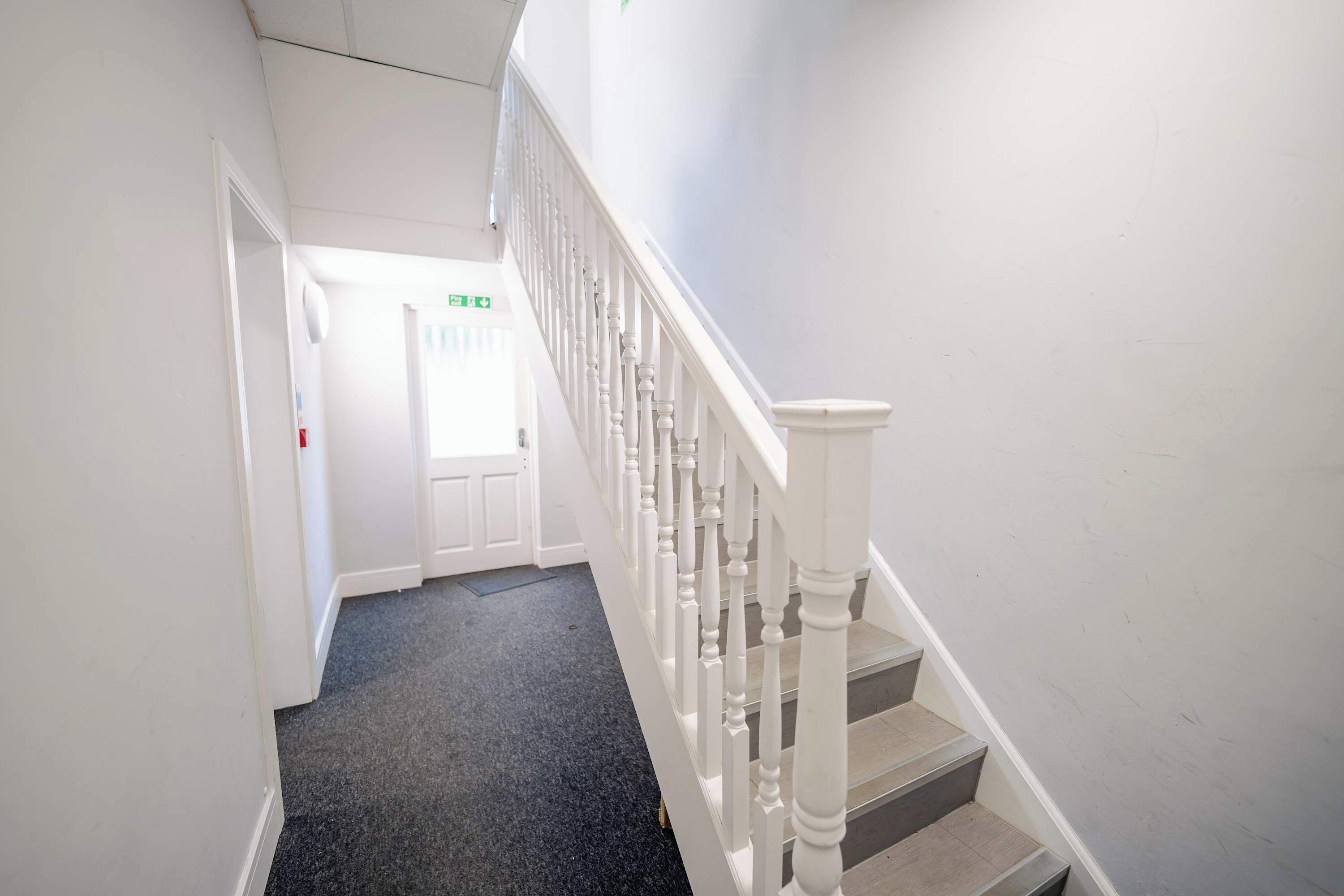A narrow hallway with a carpeted floor, white walls, and a staircase with white balusters.