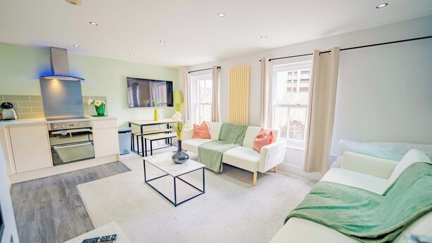 A modern living room with a sofa, coffee table, and a TV. There's a kitchen area with a stove and a dining table. - Central Gloucestershire Flat 1 Bedroom  Sleeps 6 (Gloucestershire)
