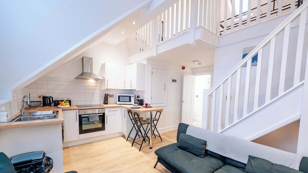A modern kitchen with white cabinetry, a dining area with a table and chairs, a sofa, and a staircase. - Elegant Cardiff Bay Flat Spacious & Comfortable (Cardiff)