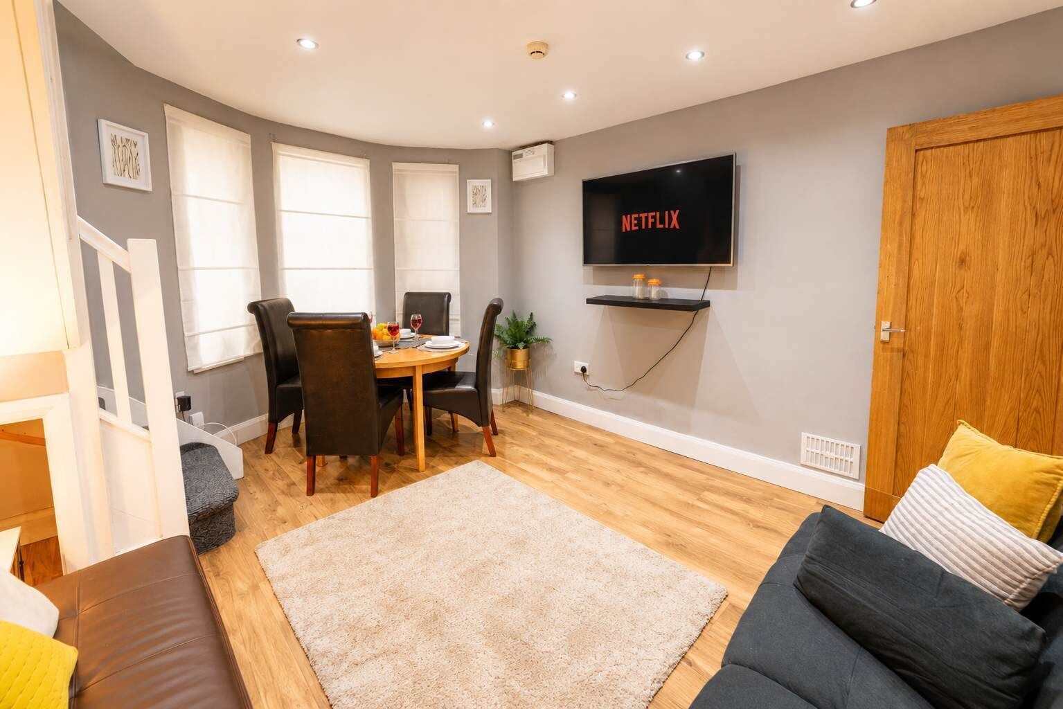 A modern living room with a dining area, a flat-screen TV mounted on the wall, and a wooden door.