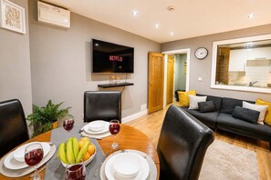 A dining area with a table set for four, a television mounted above the table, and a sofa in the background. - Cosy Modern Stay Prime - Cardiff Location - Free Parking (Cardiff)
