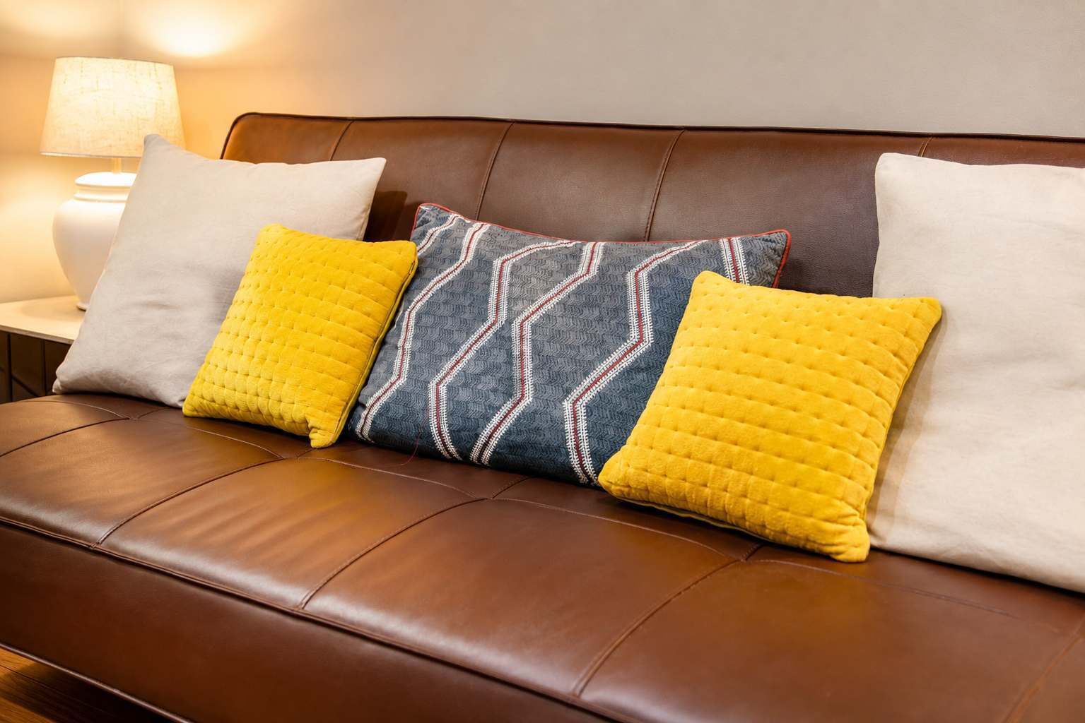 Ein braunes Ledersofa mit drei Kissen, eines gelb und zwei weiß, vor einer Wand mit einer Lampe.