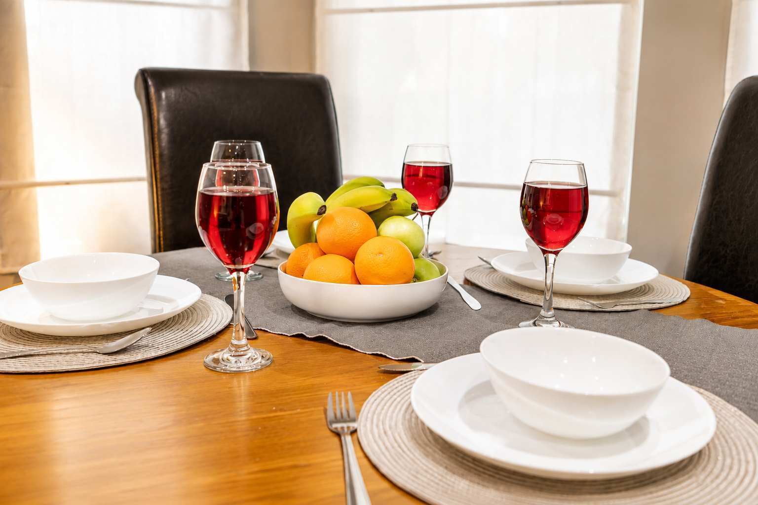 Una mesa de comedor dispuesta con platos, cuencos y cubiertos blancos, junto con un frutero y dos copas de vino tinto.