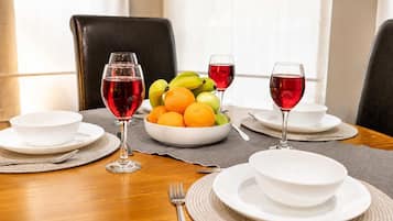 Una mesa de comedor dispuesta con platos, cuencos y cubiertos blancos, junto con un frutero y dos copas de vino tinto.