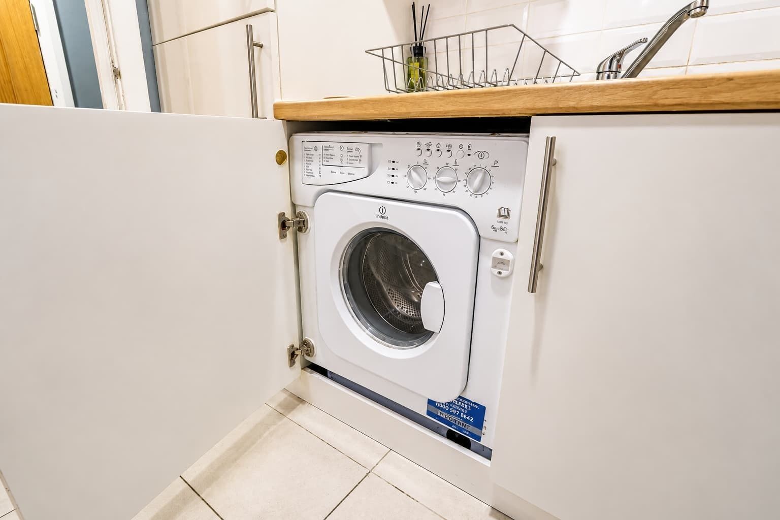 Une machine à laver compacte, de couleur blanche, à chargement frontal, installée dans une cuisine dotée d’un plan de travail en bois et d’un évier en acier inoxydable.