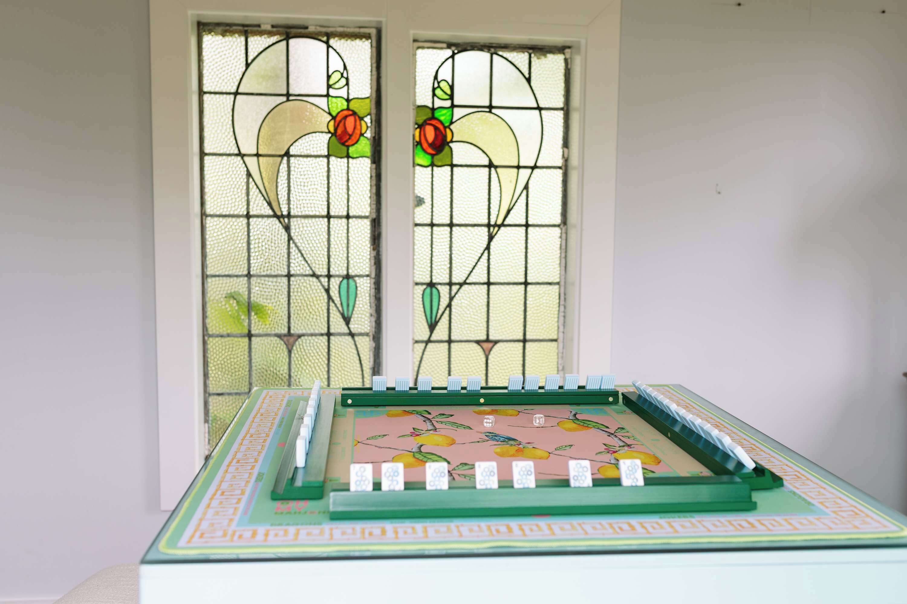 Un jeu de société est posé sur une table dont le plateau présente un motif floral, le tout étant placé devant une fenêtre ornée d’un vitrail décoratif.