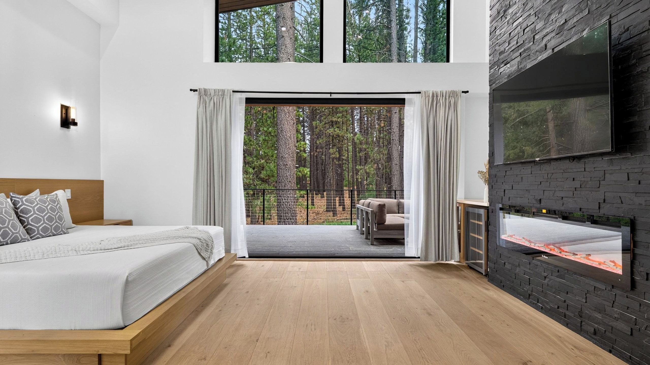 Un dormitorio con cama, suelo de madera, ventana grande, chimenea y vistas a árboles.