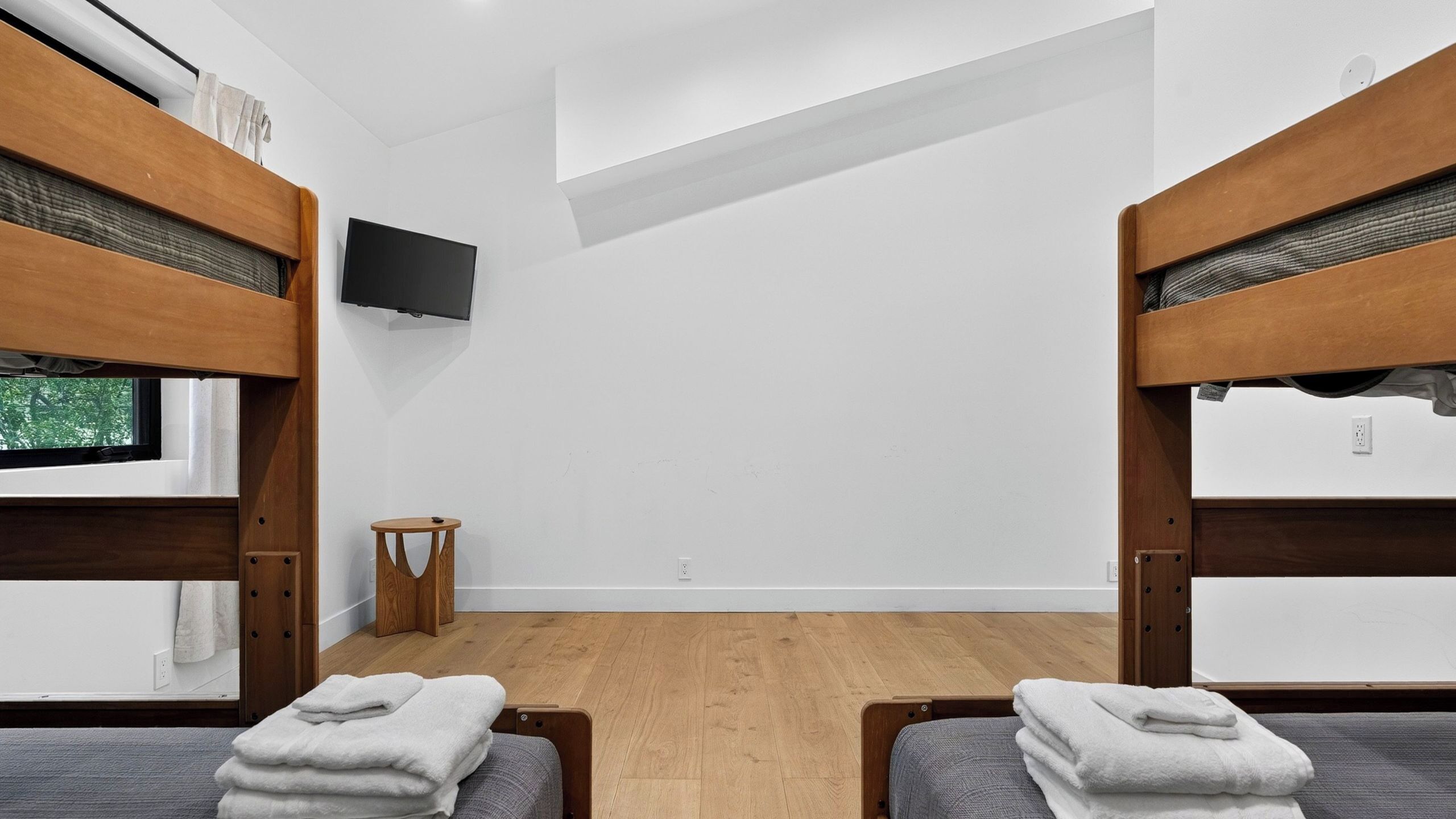 Ein Zimmer mit Etagenbetten, einem an der Wand befestigten Fernseher, einem kleinen Holzschemel und einem Fenster mit Blick auf Bäume.