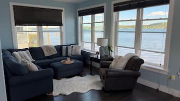Un salon avec un canapé bleu foncé, un fauteuil, un tapis blanc et de grandes fenêtres donnant sur une étendue d’eau.