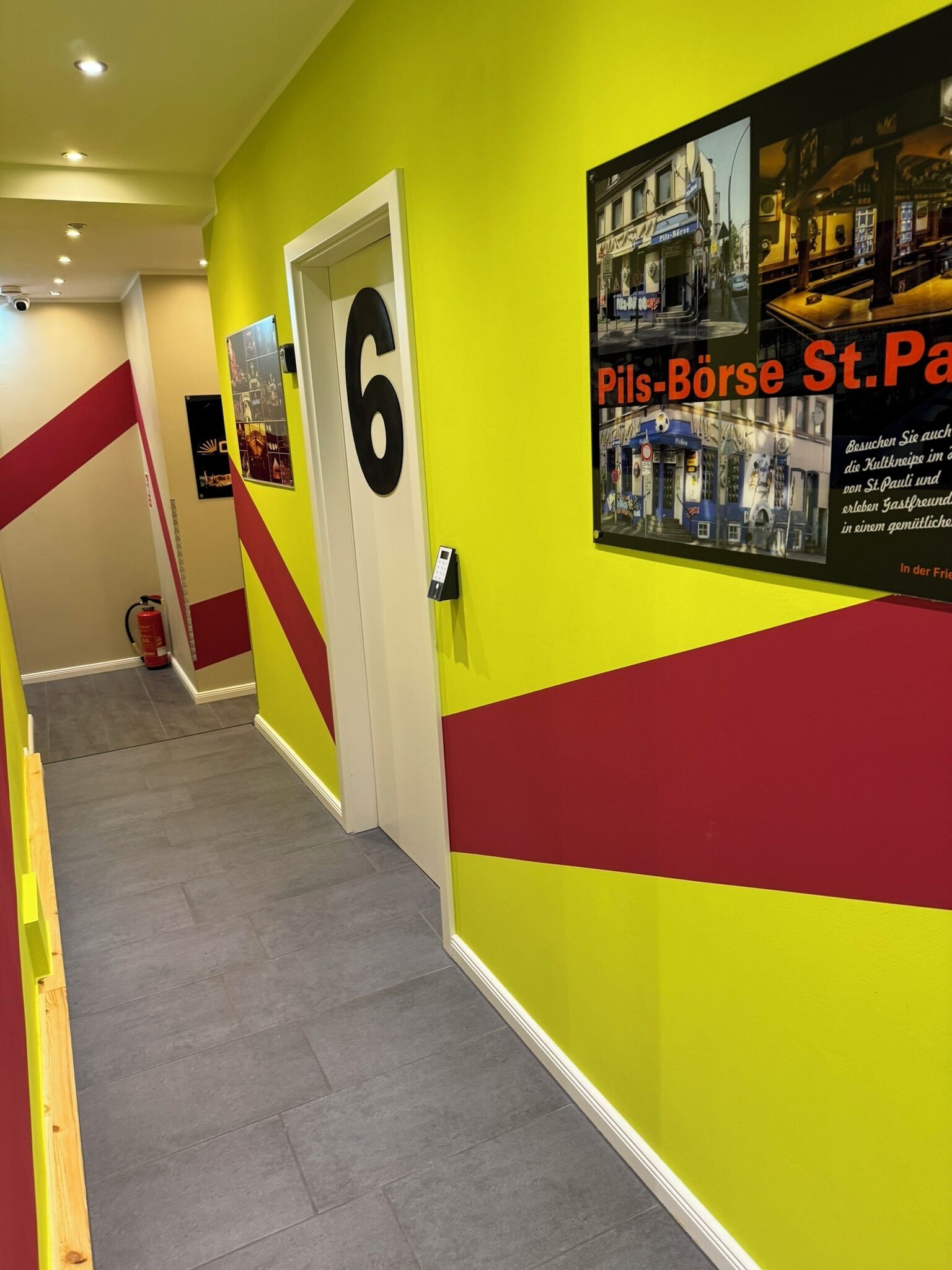 Un couloir avec un mur jaune vif, un motif rayé rouge et jaune, et une porte portant le numéro 6.