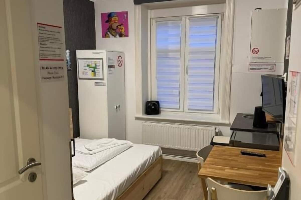 A small, well-lit room with a bed, a desk, and a window.
