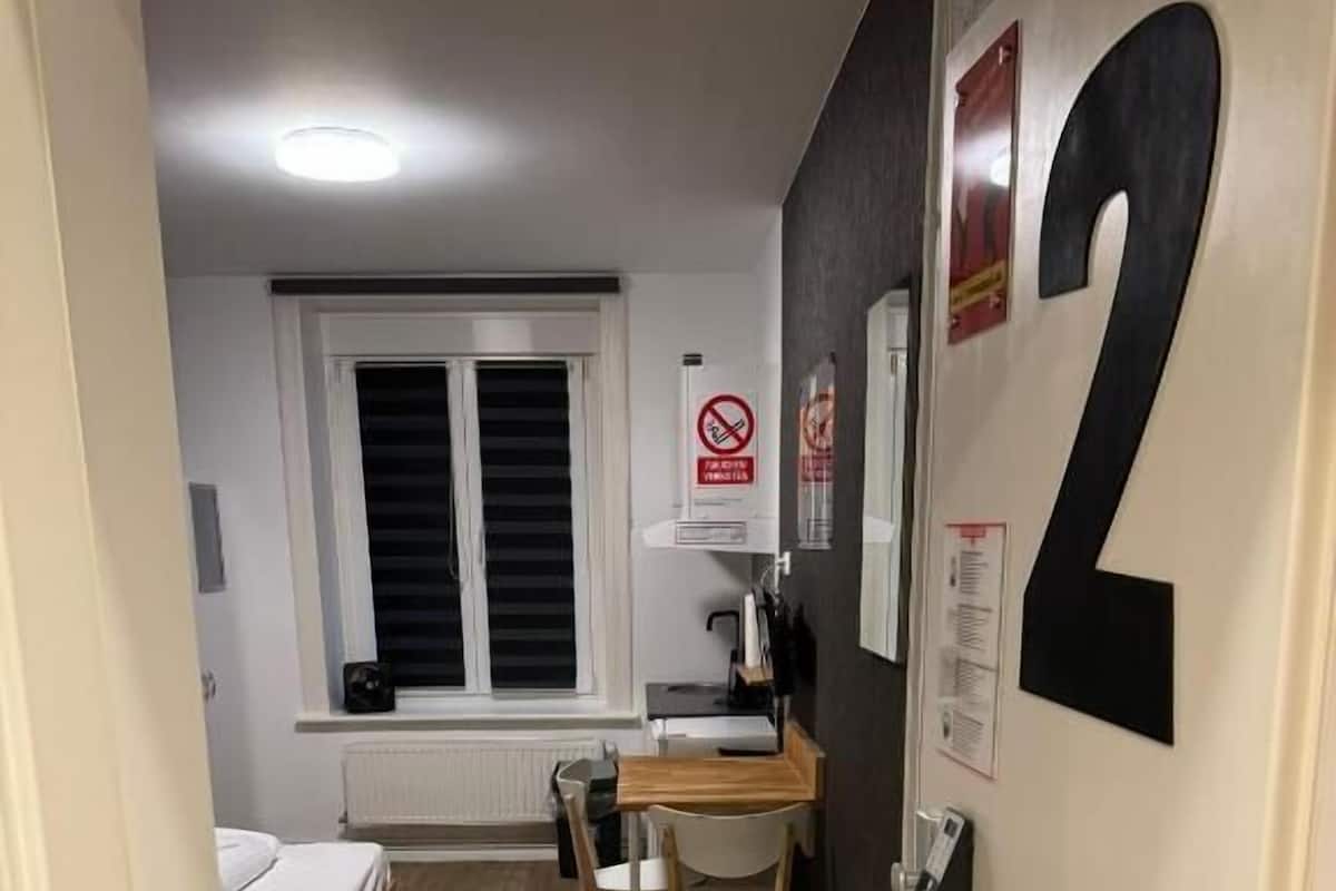 A small, compact room with a bed, a desk, and a chair. There is a window with blinds, a radiator, and a wall-mounted number '2'.