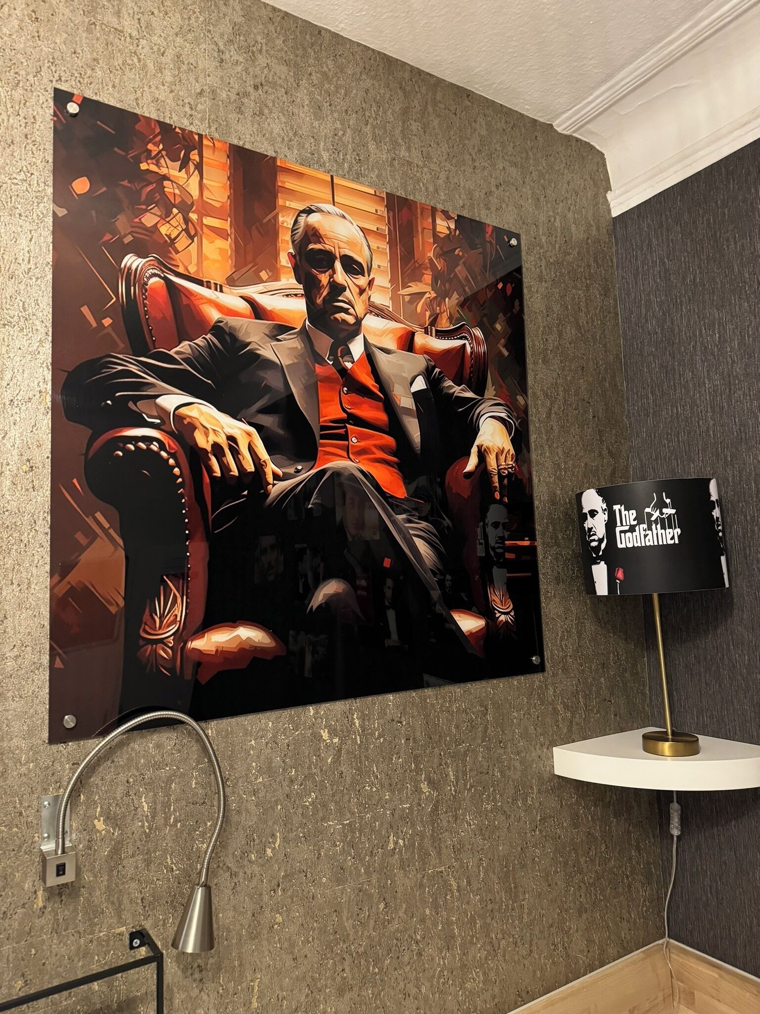 Ein großes Poster zeigt einen Mann in einem Anzug, ein kleiner Tisch mit einer Lampe und eine Wandleuchte sind zu sehen.
