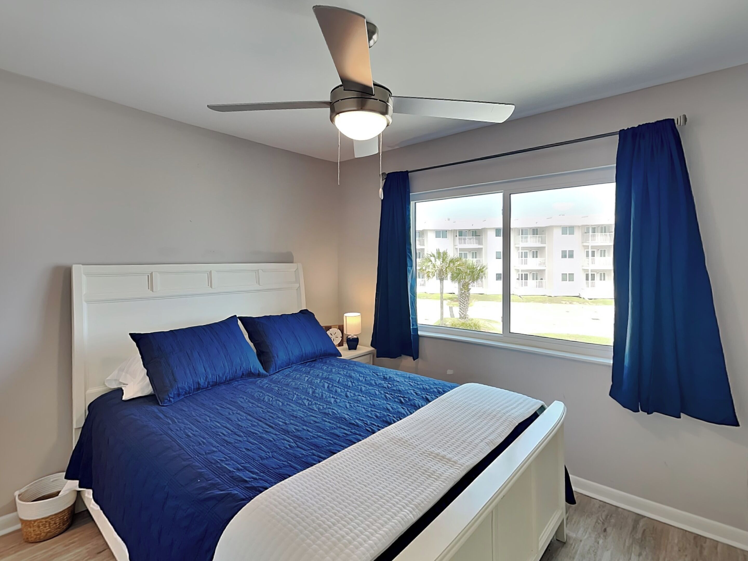 A bedroom with a bed, a ceiling fan, a window with curtains, and a view of buildings outside.