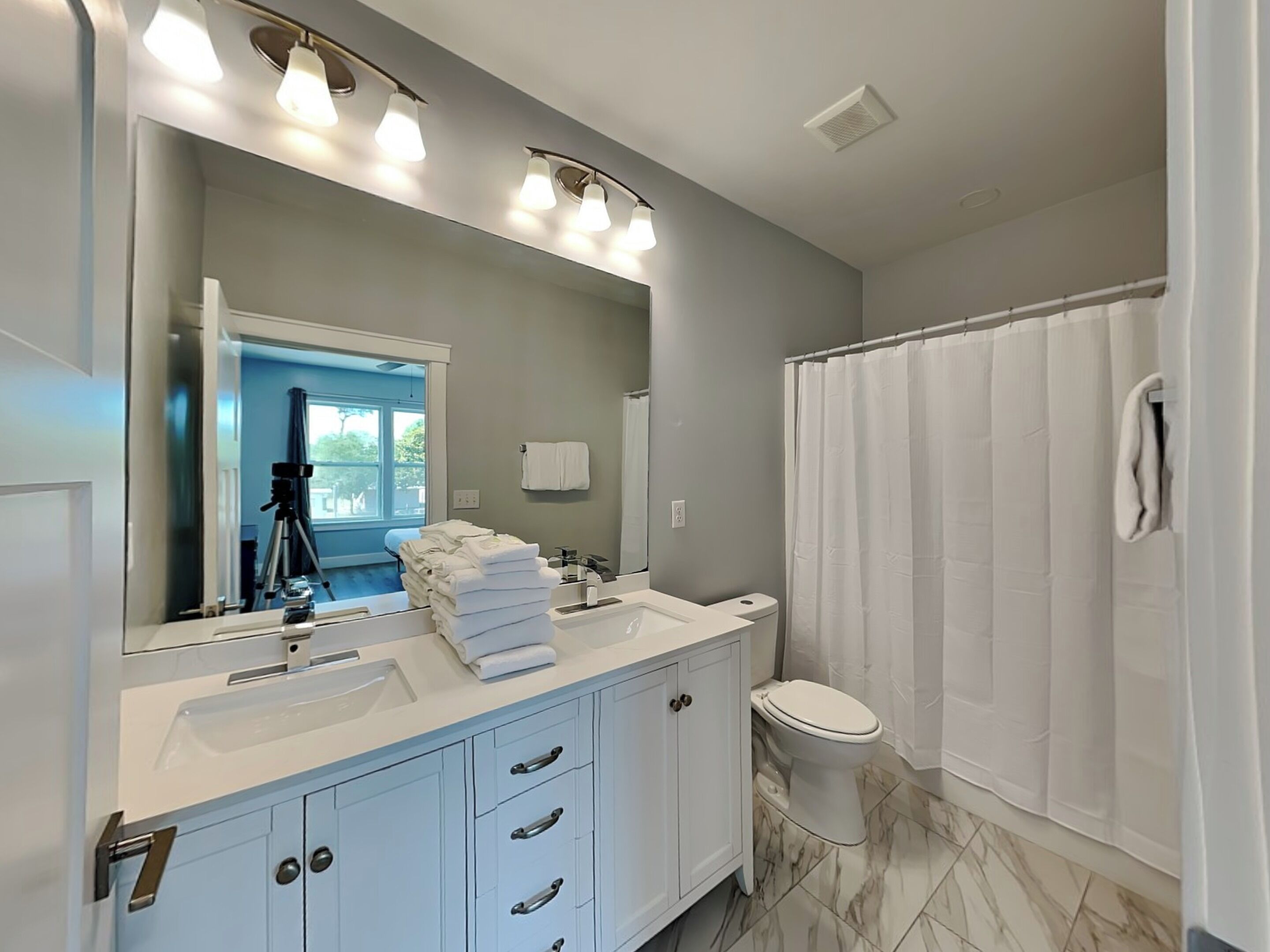 A modern bathroom with a large mirror, two sinks, a toilet, and a shower curtain.