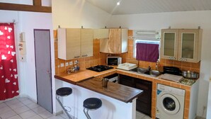 A compact kitchen with a gas stove, microwave, sink, and a washing machine. There are stools at the counter, and the kitchen has wooden cabinets and a tiled backsplash. - THE MILANOSIMPLY (Kourou)