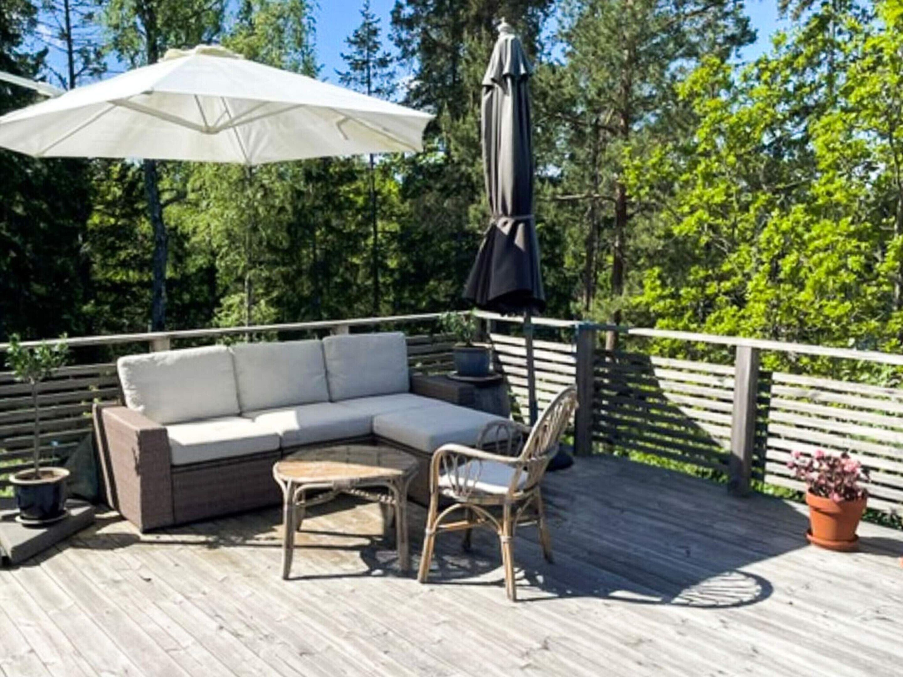 Een houten terras met een modulaire bank, twee rieten stoelen, een tafel, een witte parasol en een bloempot met plant.