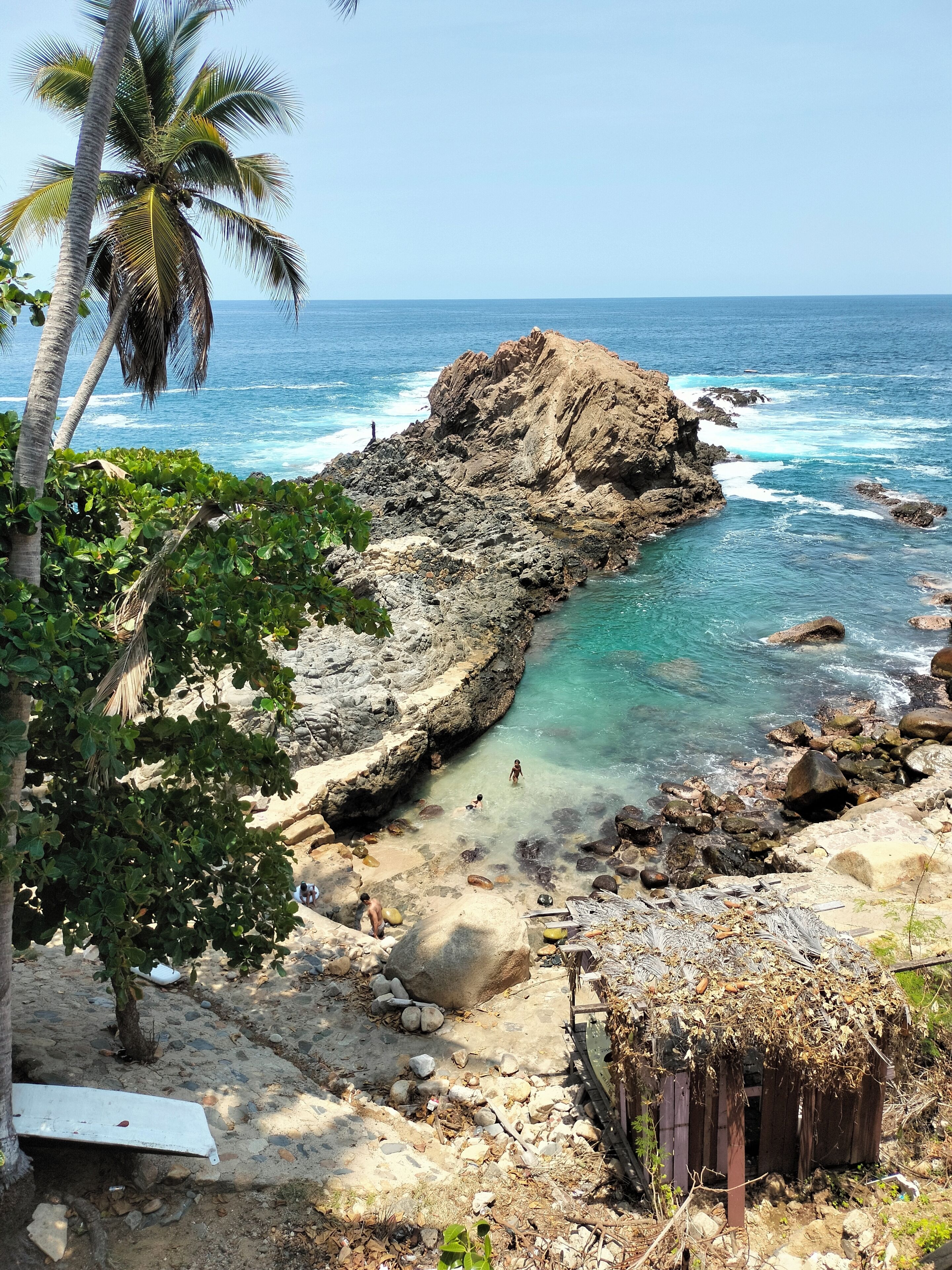 Una costa rocosa con agua azul cristalina, palmeras y una pequeña cabaña.