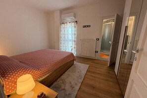 A bedroom with a bed, a nightstand, a lamp, a window with curtains, a radiator, and a shower area.