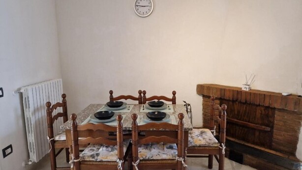 A dining area with a table set for four, wooden chairs, a clock on the wall, and a fireplace. - Tuscan Villa Retreat in San Romano (Montopoli in Val d'Arno)