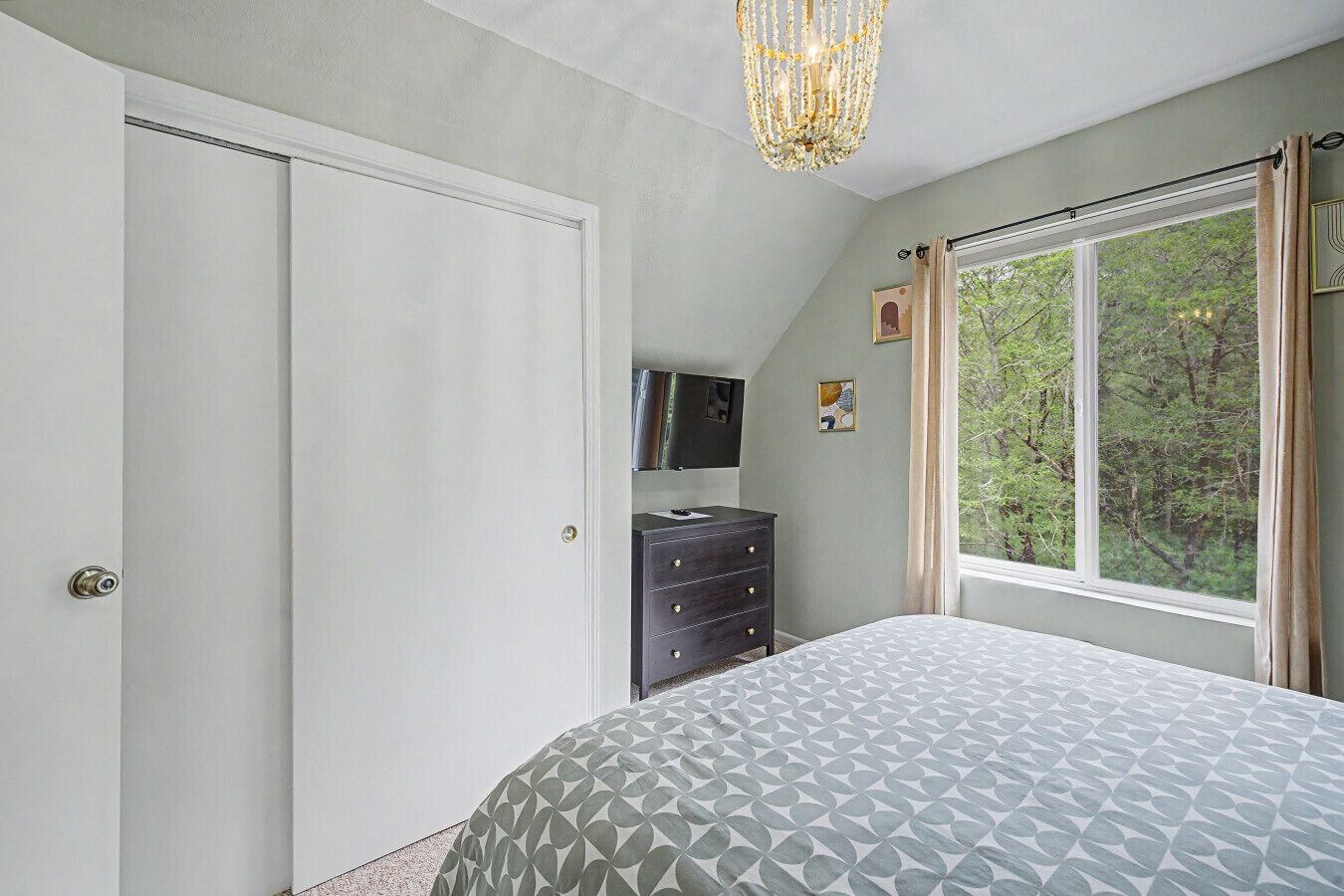 Een slaapkamer met een bed, een dressoir, een televisie en een raam met uitzicht op bomen.