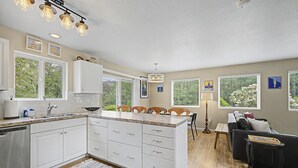 A modern kitchen with white cabinets, a granite countertop, and a dining area with a black sofa and wooden chairs. - Beach Please | Hot tub and Dog Friendly Sleeps 8 (Rockaway Beach)