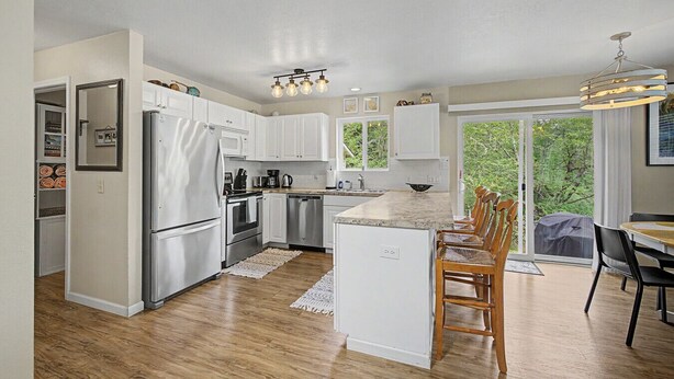 A modern kitchen with white cabinets, stainless steel appliances, a dining area with wooden chairs, and a view of greenery through large windows. - Beach Please | Hot tub and Dog Friendly Sleeps 8 (Rockaway Beach)