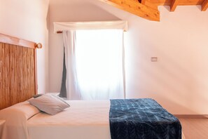 Quarto com cama, uma mesinha, janela com cortinas e teto de madeira.