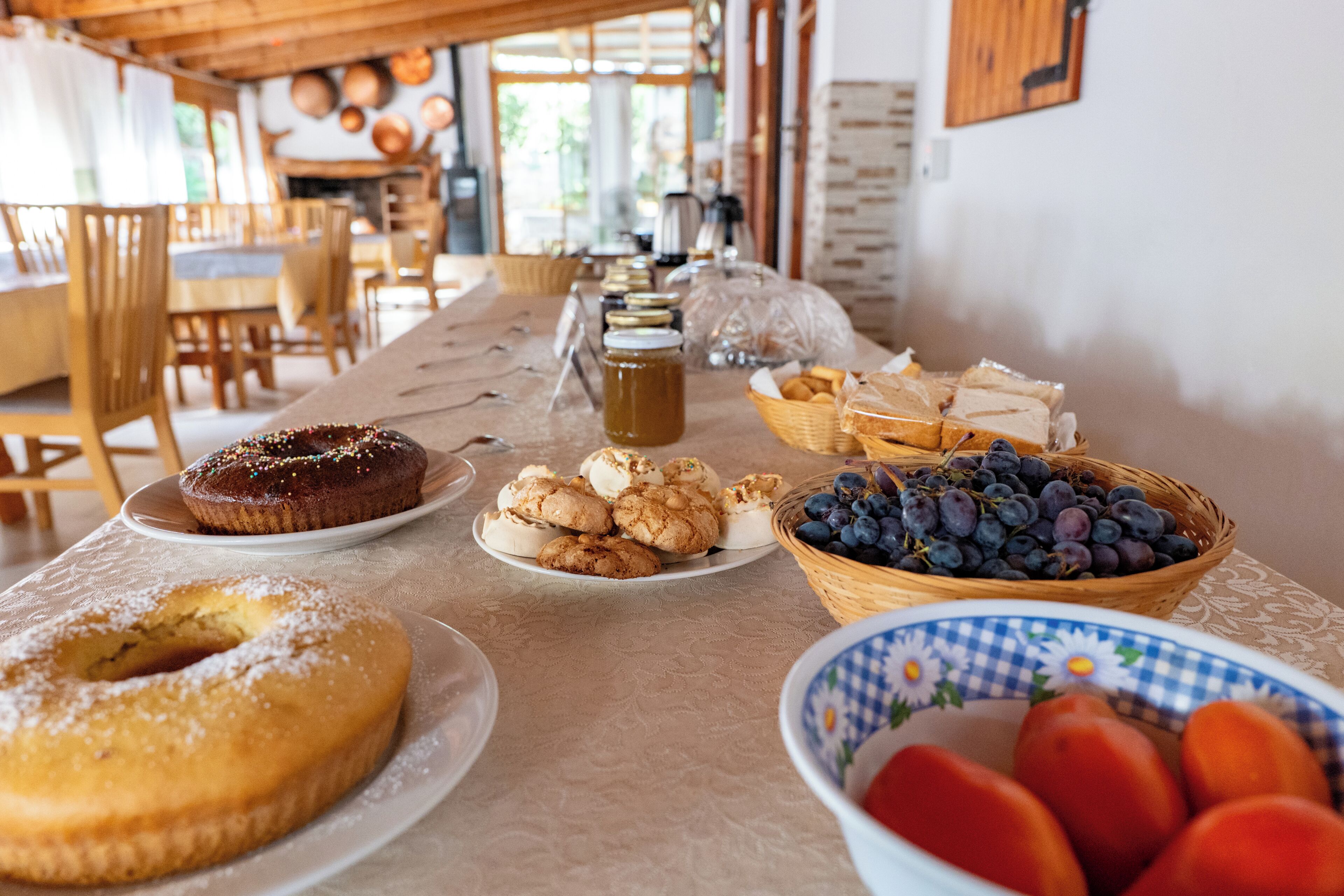 Une table dressée avec divers produits de boulangerie et des fruits, évoquant un petit-déjeuner ou une collation dans une pièce chaleureuse et bien éclairée, dotée de meubles en bois et offrant une vue sur l’extérieur.