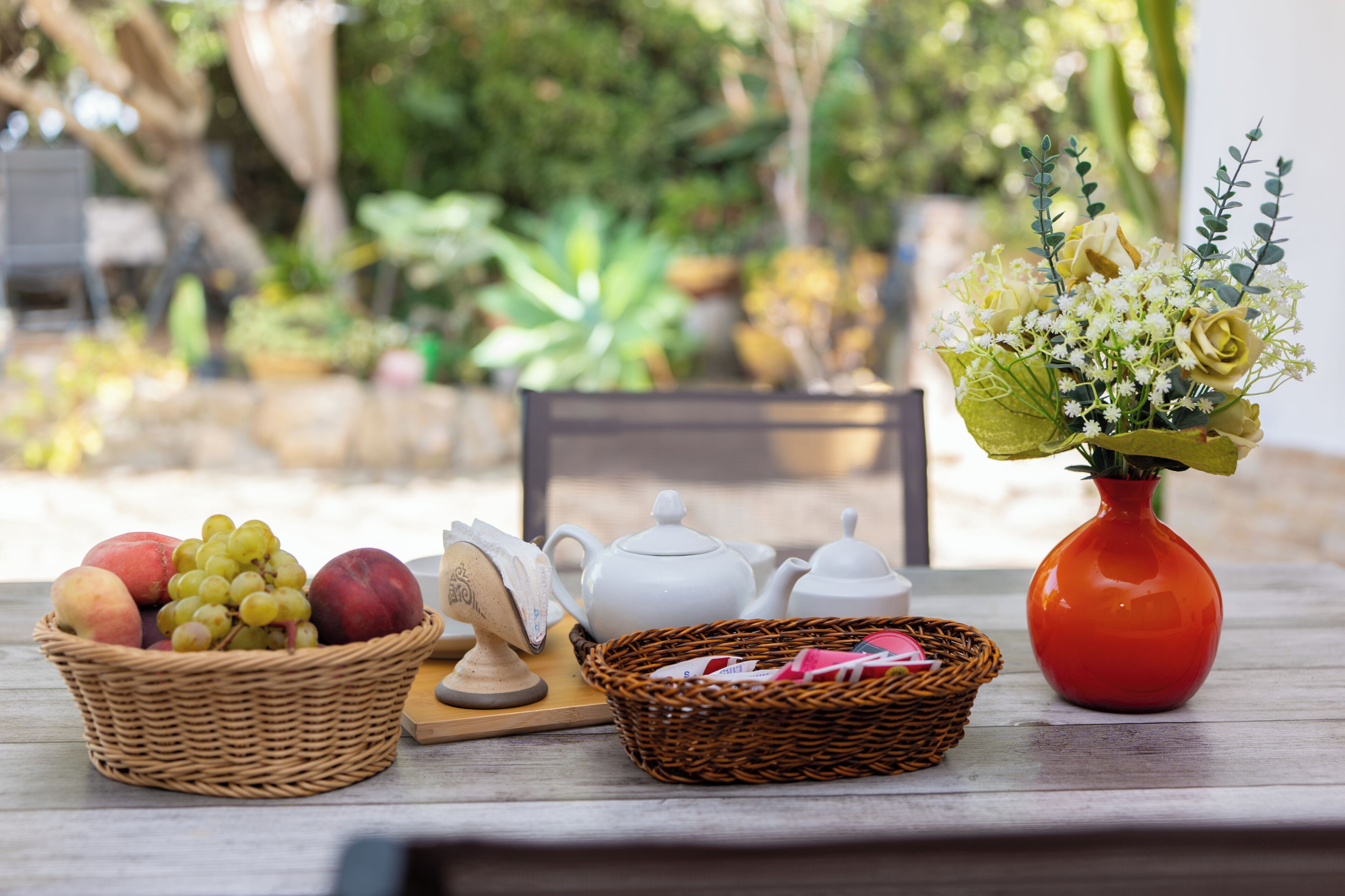 Una mesa con una cesta de fruta, un jarrón de flores y teteras blancas.