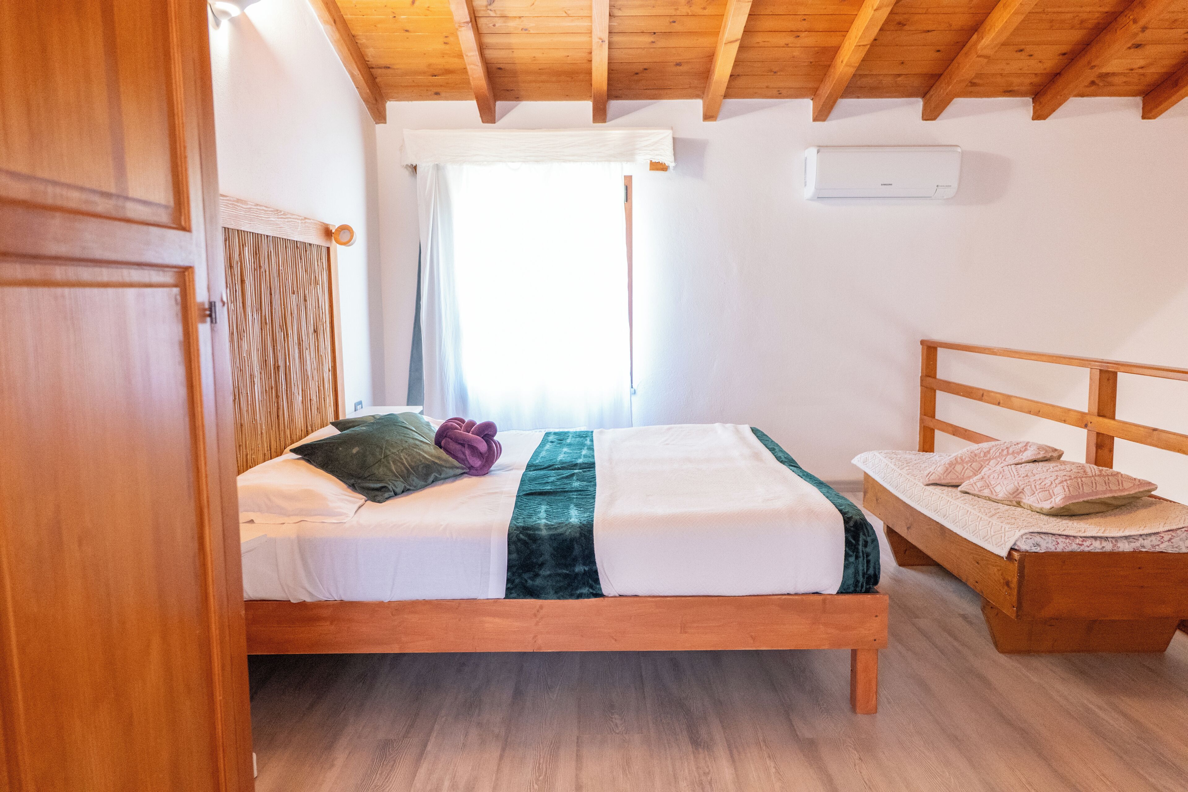 Une chambre à coucher avec un lit en bois, un banc en bois, une fenêtre avec des rideaux et un ventilateur de plafond.
