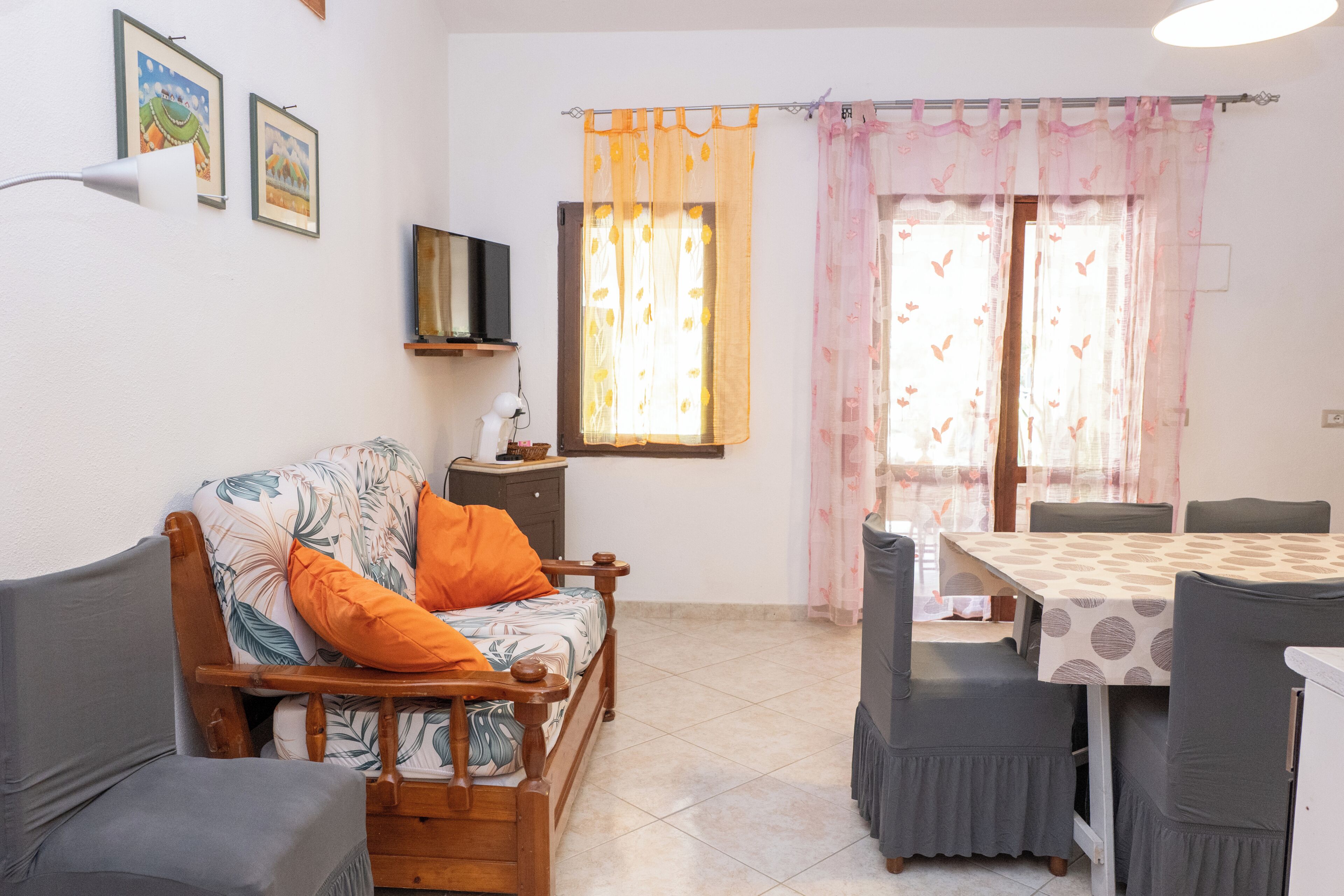 A furnished living room with a sofa, a dining table with chairs, a television on a stand, and a window with curtains.
