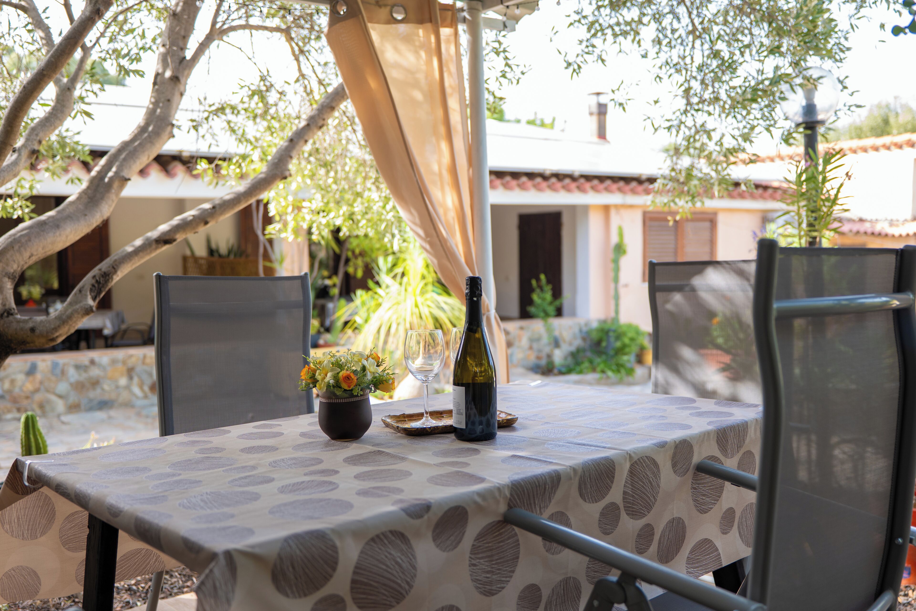 An outdoor dining area with a table set for two, surrounded by chairs, a potted plant, and a bottle of wine.