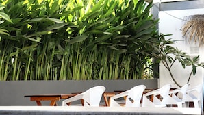 Une piscine entourée de hautes plantes vertes, avec des chaises en plastique blanc et une table en bois.