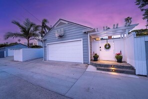 Eine Garage mit Wohntyp-Architektur, einer weißen Tür, einem überdachten Balkon und einem kleinen Gartenbereich mit Topfpflanzen.