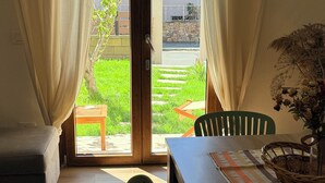 A room with a wooden floor, a table with chairs, a bench, and a view through a glass door to an outdoor area with greenery and a building. - Su Entu Suite Free Parking 5 min from the sea (Giba)