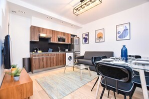 Eine moderne Küche mit Holzausstattung, einem Einbauofen und einem Kühlschrank. Es gibt einen Essbereich mit einem Glastisch und Stühlen, einer Couch und Kunstwerken an den Wänden.
