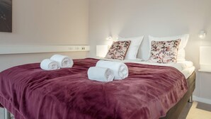 Ein ordentlich bezogenes Bett mit einer dunkelroten Decke, weißen Kissen und zwei aufgerollten Handtüchern.