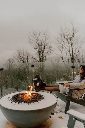 A person relaxing by a fire pit with a bench, surrounded by a snowy landscape and bare trees.