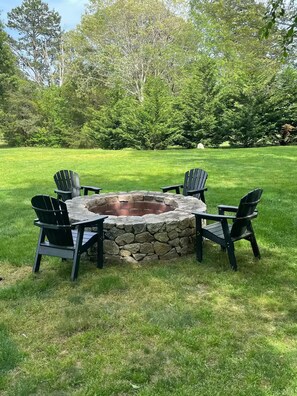 A stone fire pit surrounded by four Adirondack chairs in a grassy outdoor area.