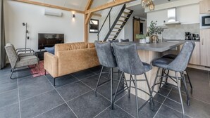 Een ruime woonkamer met een hoge plafond, een bank, barstoelen, een televisie en een keukenhoek met een trap.