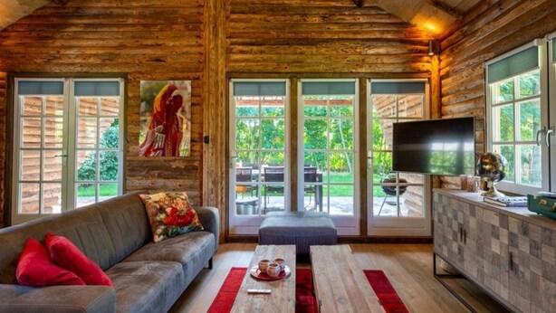 Een interieur van een blokhut met een bank, een houten tafel, een televisie en uitzicht op de omgeving.
