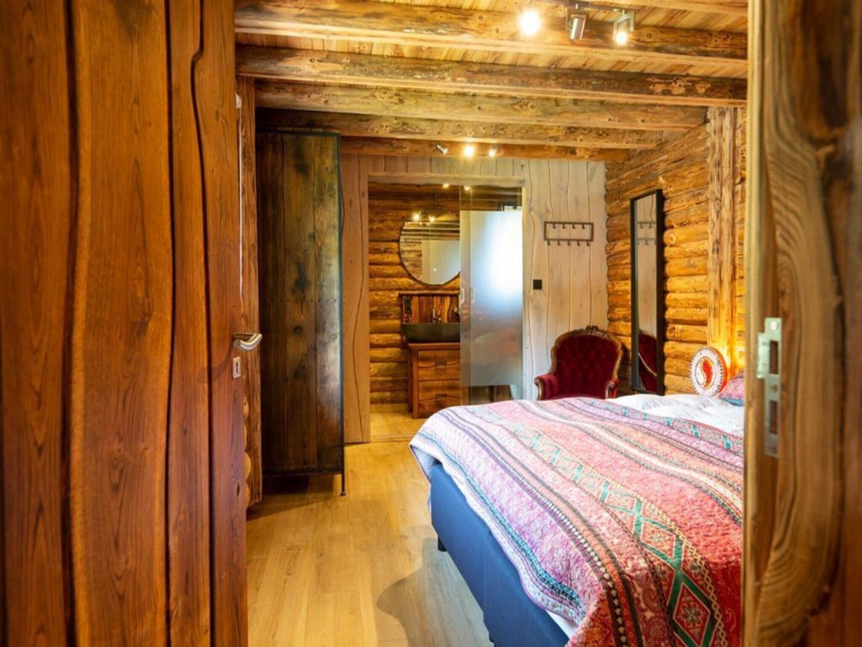 Un dormitorio de estilo cabaña de troncos con techo de madera, una cama con colcha estampada, una silla roja y un aparador de madera.