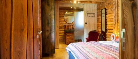 Un dormitorio de estilo cabaña de troncos con techo de madera, una cama con colcha estampada, una silla roja y un aparador de madera.