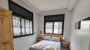 A bedroom with a bed, two nightstands, a wooden wardrobe, and a window with a metal gate. - Villa Aronora Private Pool & BBQ (Haddada)
